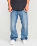 A person is shown from the shoulders down, wearing a black patterned shirt, Volcom Men’s Pipe Dreams Denim Pants in blue, and white sneakers, standing against a plain light background.