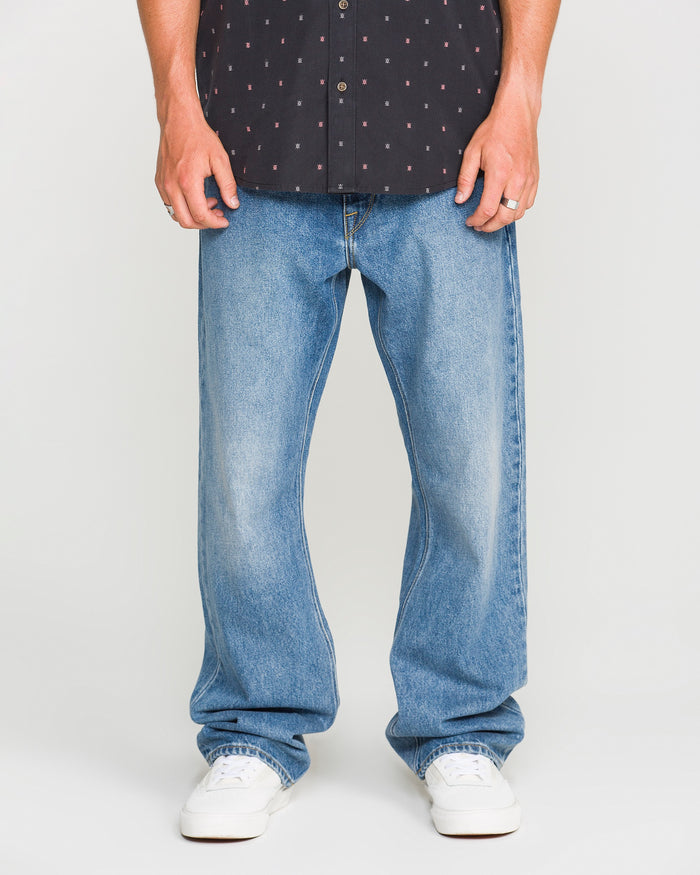 A person is shown from the shoulders down, wearing a black patterned shirt, Volcom Men’s Pipe Dreams Denim Pants in blue, and white sneakers, standing against a plain light background.