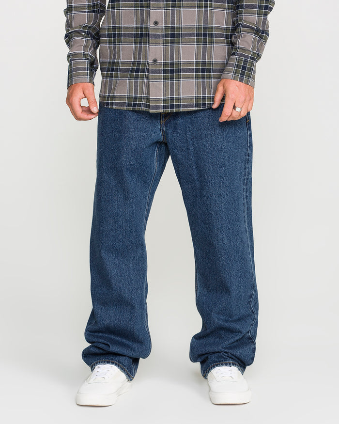 A person wearing a gray plaid shirt, Volcom Men’s Pipe Dreams Denim Pants in a relaxed straight fit with rigid 100% cotton denim, and white sneakers stands against a plain background. Only the lower half of their body is visible.