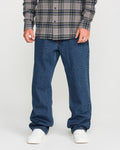 A person wearing a gray plaid shirt, Volcom Men’s Pipe Dreams Denim Pants in a relaxed straight fit with rigid 100% cotton denim, and white sneakers stands against a plain background. Only the lower half of their body is visible.