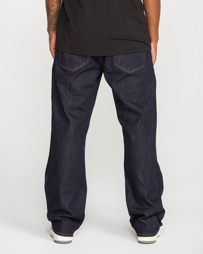 A person wearing a black t-shirt and loose dark blue jeans stands facing away, hands at their sides. They are wearing Volcom Men’s Pipe Dreams Denim Pants, crafted from indigo stretch denim with a relaxed straight leg fit, back pockets, and visible stitching. The look is completed with white sneakers.