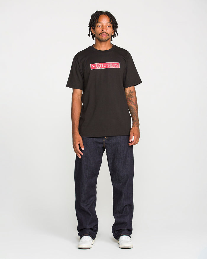 A man with short dreadlocks and tattoos on his left arm stands facing forward against a plain white background, wearing a black Volcom T-shirt, white shoes, and Volcom Men’s Pipe Dreams Denim Pants in indigo stretch denim with a relaxed straight leg fit.