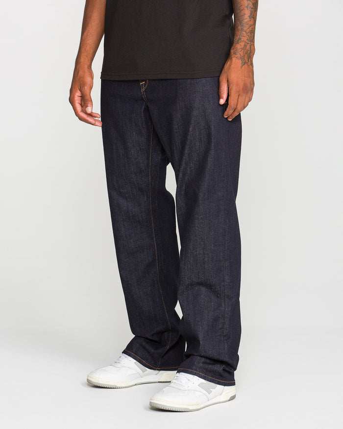 Wearing Volcom’s Men’s Pipe Dreams Denim Pants in indigo stretch denim, paired with a black t-shirt and white sneakers, an individual stands against a plain background. Only the lower half of the body is visible.