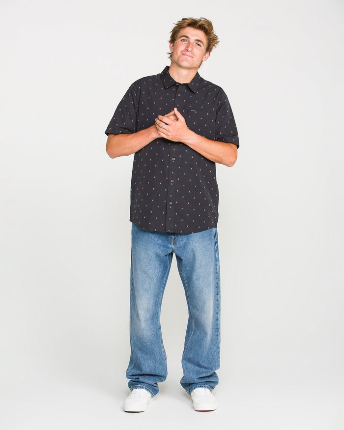 A person stands against a white background, wearing a black short-sleeve button-up shirt with a small pattern, Volcom Men’s Pipe Dreams Denim Pants in a relaxed straight fit vintage-inspired blue denim, and white sneakers, with hands clasped over their chest and a slight smile.