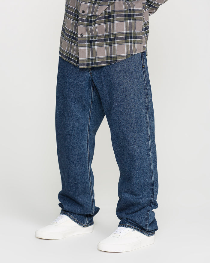 A person wearing a plaid shirt, Volcom Men’s Pipe Dreams Denim Pants featuring a relaxed straight fit crafted from 100% cotton denim, and white sneakers stands against a plain background. Only the lower half of the body is visible.