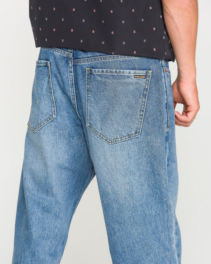 A person shown from the waist down and back is wearing Volcom Men’s Pipe Dreams Denim Pants—loose-fitting, vintage-inspired denim blue jeans with large back pockets and a small Volcom label—paired with a black patterned shirt.