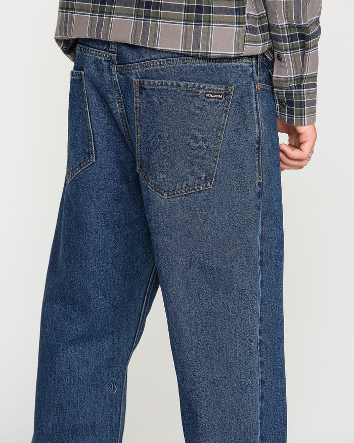 A person wearing a plaid shirt and Volcom Men’s Pipe Dreams Denim Pants in a relaxed straight fit dark indigo color stands with their back to the camera, showcasing the back pocket and part of the leg crafted from rigid 100% cotton denim.