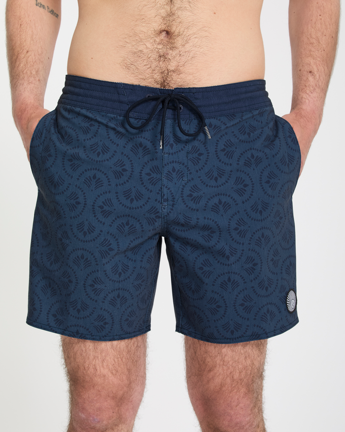 A man wears the Volcom Men's Psyche Cotton Stoney 17 swim trunks—dark blue with a pattern and drawstring—hands in pockets, standing against a plain white background.