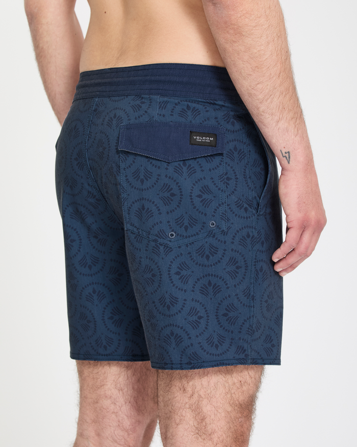 A person stands with their back to the camera in Volcom's Men's Psyche Cotton Stoney 17 shorts, featuring a navy blue pattern, dark waistband, right-side flap pocket, and a visible tattoo on their left forearm.