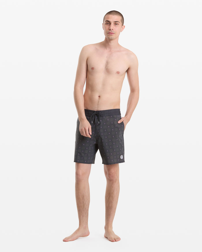 A young man with short hair stands barefoot against a white backdrop, wearing Volcom's Men's Psyche Cotton Stoney 17 swim trunks. He is shirtless, has one hand in his pocket, and displays a neutral expression.