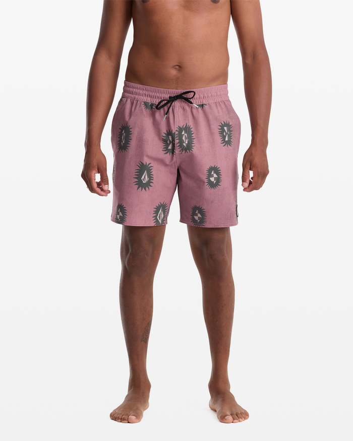 A person models Volcom's Men's Psyche Cotton Trunk 17 swim shorts, featuring a mauve color with a black abstract sunburst pattern, shown from shoulders to feet against a white background.