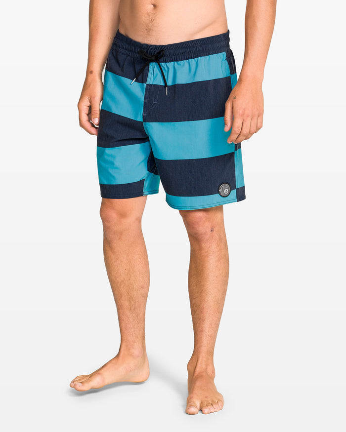 A person stands barefoot against a plain white background wearing Volcom Men's Psyche Cotton 17" Trunk swim shorts with a drawstring waist and side pocket.