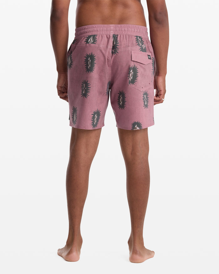 A person stands barefoot facing away, wearing Volcom Men's Psyche Cotton Trunk 17 shorts in pink with a black cactus pattern, featuring an elastic waistband and a back pocket.