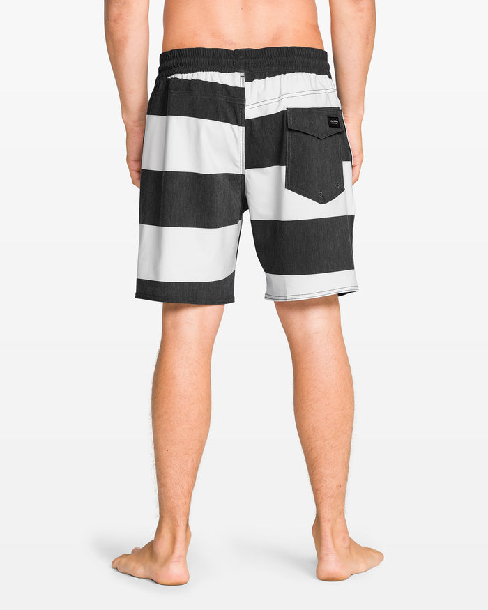A man stands barefoot, facing away, wearing Volcom Men's Psyche Cotton 17" Trunk—black and white horizontal stripes, elastic waistband, back pocket.