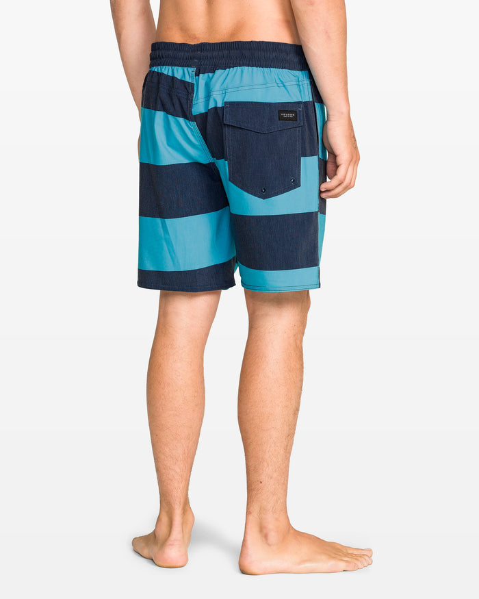 A person stands barefoot, facing away, wearing Volcom Men's Psyche Cotton 17" Trunk—swim shorts with blue and navy stripes, elastic waistband, back pocket, and 17-inch outseam. The background is plain white.
