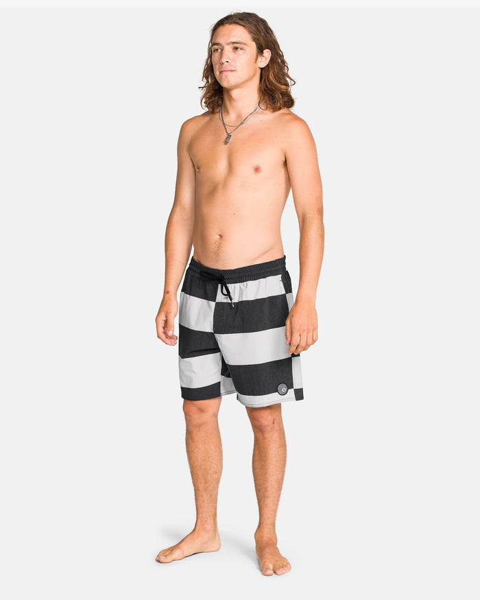 A young man with long brown hair stands barefoot on a white background, wearing Volcom Men's Psyche Cotton 17" Trunk swimwear and a necklace. He is shirtless and looking slightly to the side.
