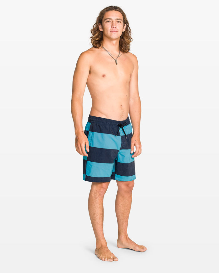 A young man with long brown hair stands barefoot on a plain background, wearing the Volcom Men's Psyche Cotton 17" Trunk—blue and navy striped swim shorts. He is shirtless, facing slightly to the side, looking at the camera and wearing a necklace.