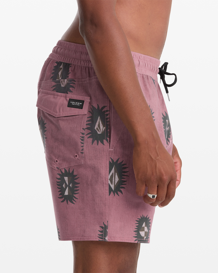 A person shown from the waist down, side view, wearing Volcom Men's Psyche Cotton Trunk 17 in mauve with black and white geometric sun patterns, a black drawstring, and a side pocket featuring a rectangular logo patch.