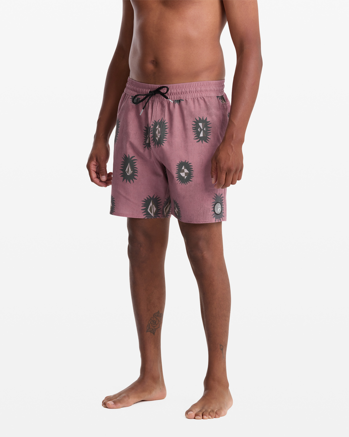 A man stands barefoot wearing Volcom Men's Psyche Cotton Trunk 17 swim trunks in mauve with a black abstract sunburst pattern, black drawstring, and side pockets. His upper body and head are not shown; the background is plain white.