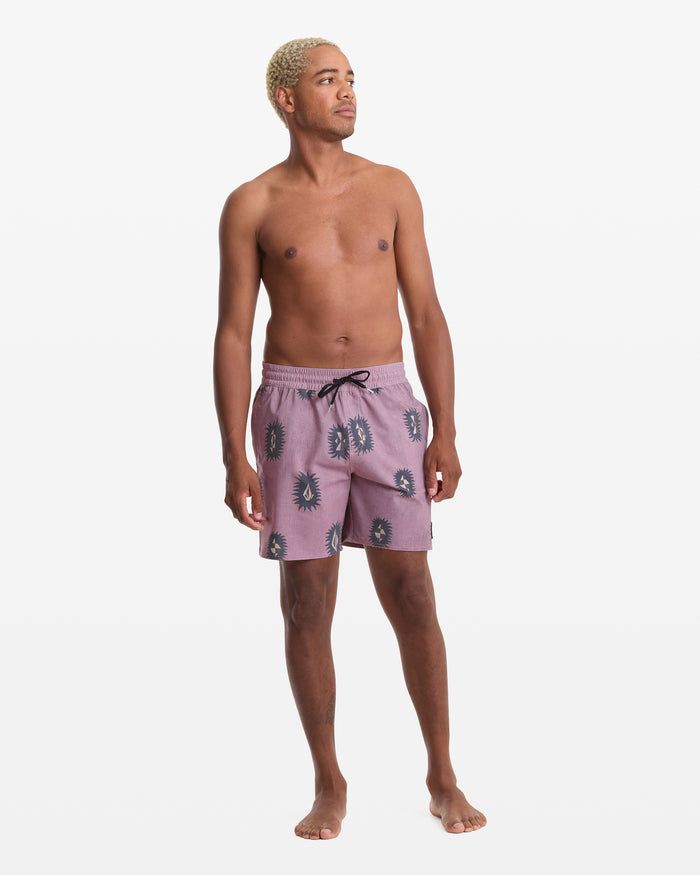 A shirtless man with short blond hair stands barefoot against a white background, wearing the Volcom Men's Psyche Cotton Trunk 17—mauve swim trunks with a black and white abstract face print and black drawstring—looking slightly to his left.