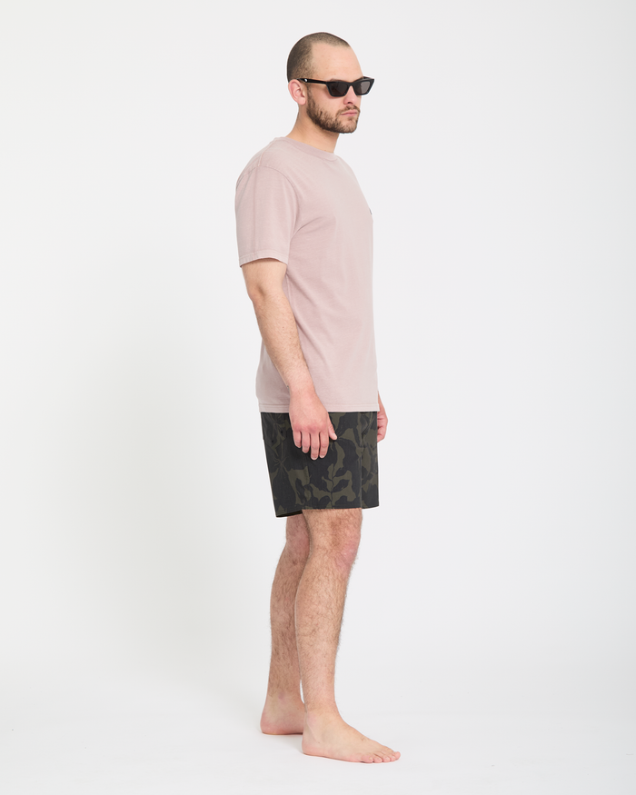 A man stands barefoot in profile against a white background, wearing black sunglasses, a pale pink t-shirt, and Volcom Men's Psyche Cotton Trunk 17 shorts.