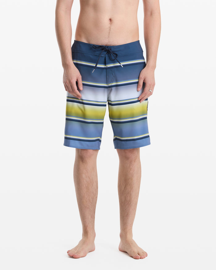 A person stands barefoot, shown from the waist down against a plain white background, wearing Volcom Men's Quarta Dissolve Mod 20 swim trunks featuring blue, white, and yellow stripes with a drawstring waist.