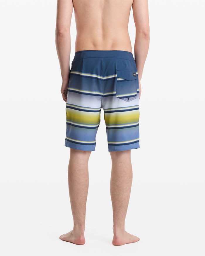 A man stands barefoot with his back to the camera, wearing Volcom Men's Quarta Dissolve Mod 20 blue board shorts featuring horizontal white, yellow, and green stripes and a right-side pocket.