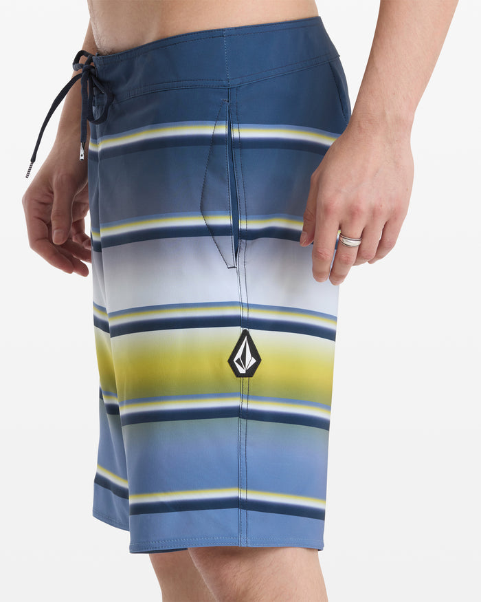 A person wearing Volcom Men's Quarta Dissolve Mod 20 board shorts featuring blue, white, and yellow stripes, a drawstring waist, and a small black and white left leg logo. Only the torso and upper legs are shown on a white background.