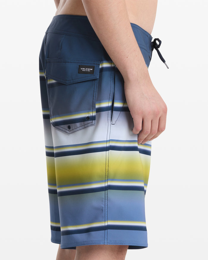 A person shown from the side wears Volcom Men's Quarta Dissolve Mod 20 board shorts featuring blue, white, yellow, and green stripes, a back pocket, and a black drawstring.