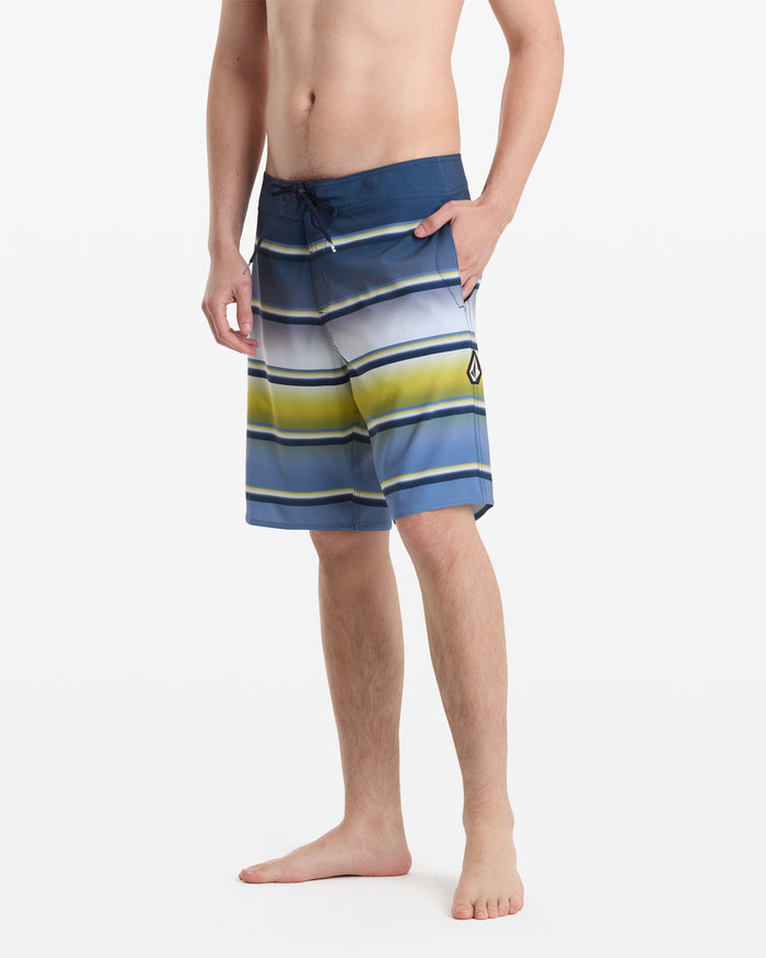 A barefoot man wears Volcom Men's Quarta Dissolve Mod 20 board shorts with blue, white, yellow, and gray stripes, a drawstring waist, and a small logo on the left leg. His torso is bare, and one hand rests on his hip.