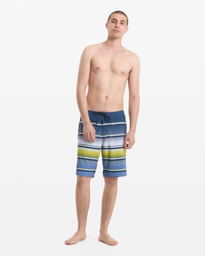 A young man stands barefoot against a white background, wearing Volcom Men's Quarta Dissolve Mod 20 board shorts with blue, green, and white stripes. He is shirtless with a relaxed posture and neutral expression.