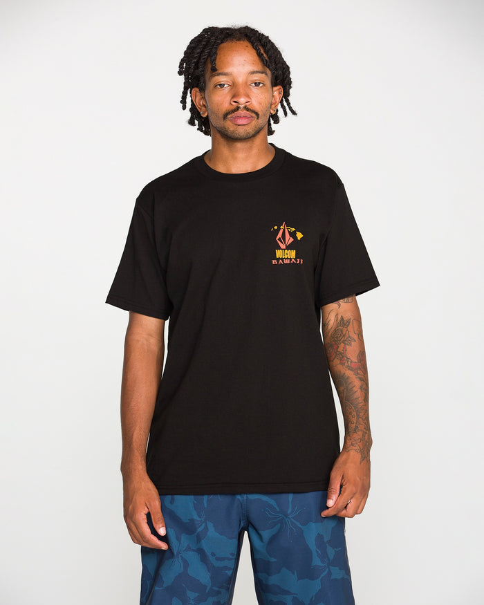 A man with short dreadlocks and tattoos on his left arm wears the Volcom Men's Refractor Short Sleeve Tee, a classic fit made from premium ring spun cotton with a small, colorful soft-hand screen print graphic on the chest. He pairs it with blue camo shorts while standing against a plain white background.