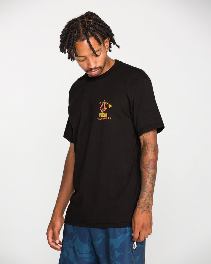 A man with braided hair wears the Volcom Men's Refractor Short Sleeve Tee, a black graphic T-shirt crafted from premium ring spun cotton and featuring a soft-hand screen print for extra comfort. He pairs it with blue patterned shorts and stands against a white background, looking down with one hand by his side.