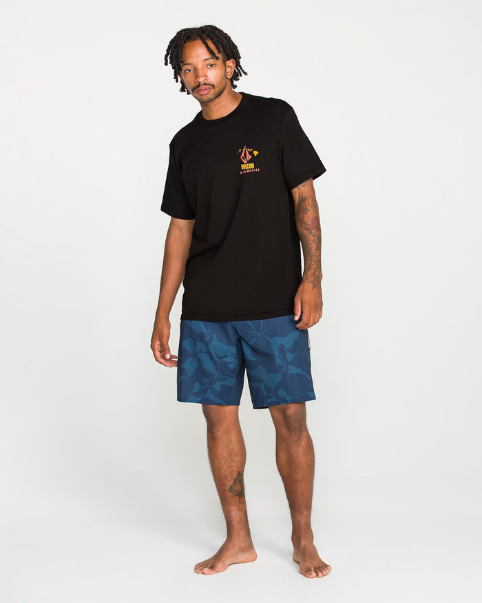 A man stands barefoot on a white background, wearing the Volcom Men's Refractor Short Sleeve Tee, a classic fit made from premium ring spun cotton with a small yellow graphic and soft-hand screen print on the chest, paired with blue floral-patterned shorts.