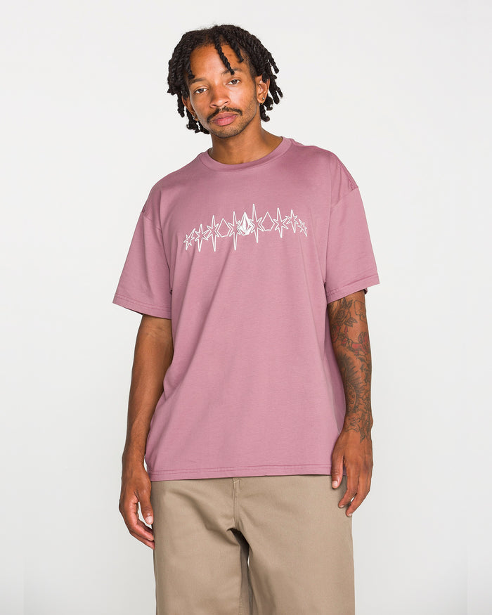 A man wearing the Volcom Men's Repeater Short Sleeve Tee in mauve, featuring a white abstract graphic across the chest and paired with beige pants, stands against a plain white background. This loose fit crew neck tee is crafted from heavyweight ringspun cotton for exceptional comfort and durability.