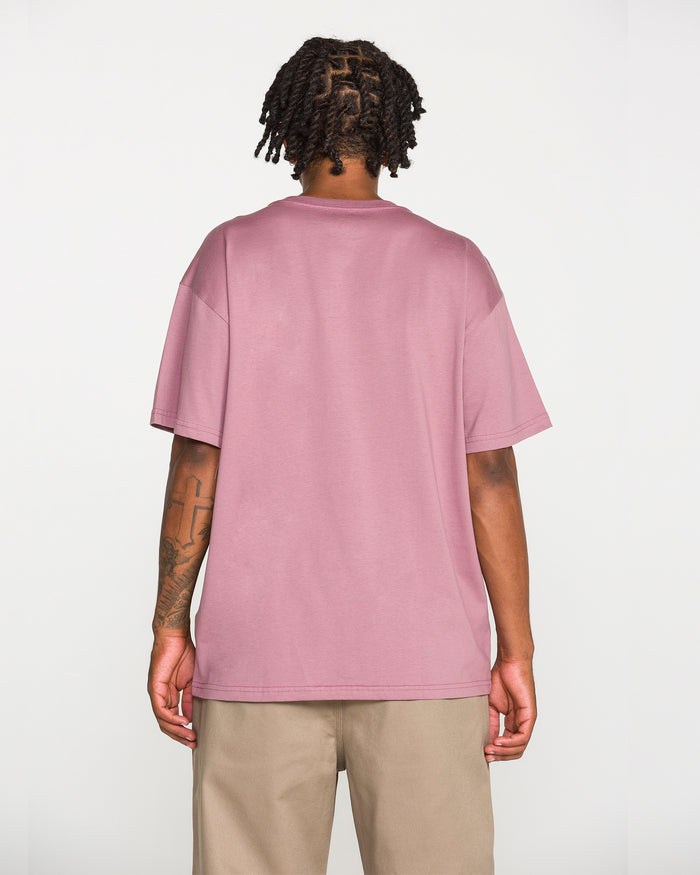 A person with short twisted hair stands facing away from the camera, wearing Volcom's Men's Repeater Short Sleeve Tee in mauve—a loose fit crafted from heavyweight ringspun cotton—paired with beige pants, showcasing the back of the shirt.
