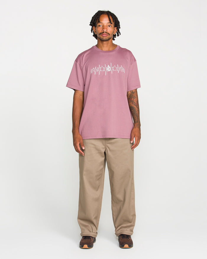 A man with short dreadlocks wears the Volcom Men's Repeater Short Sleeve Tee in a mauve hue, featuring a loose fit and crafted from heavyweight ringspun cotton. He pairs it with beige pants and brown shoes, standing facing forward against a plain white background with a neutral expression.