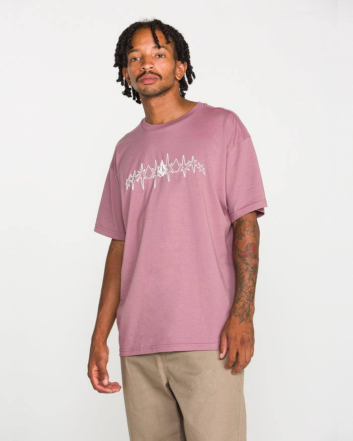 A man with short dreadlocks wears the Volcom Men's Repeater Short Sleeve Tee—a mauve loose-fit t-shirt with a white abstract design—paired with beige pants, standing against a plain white background.