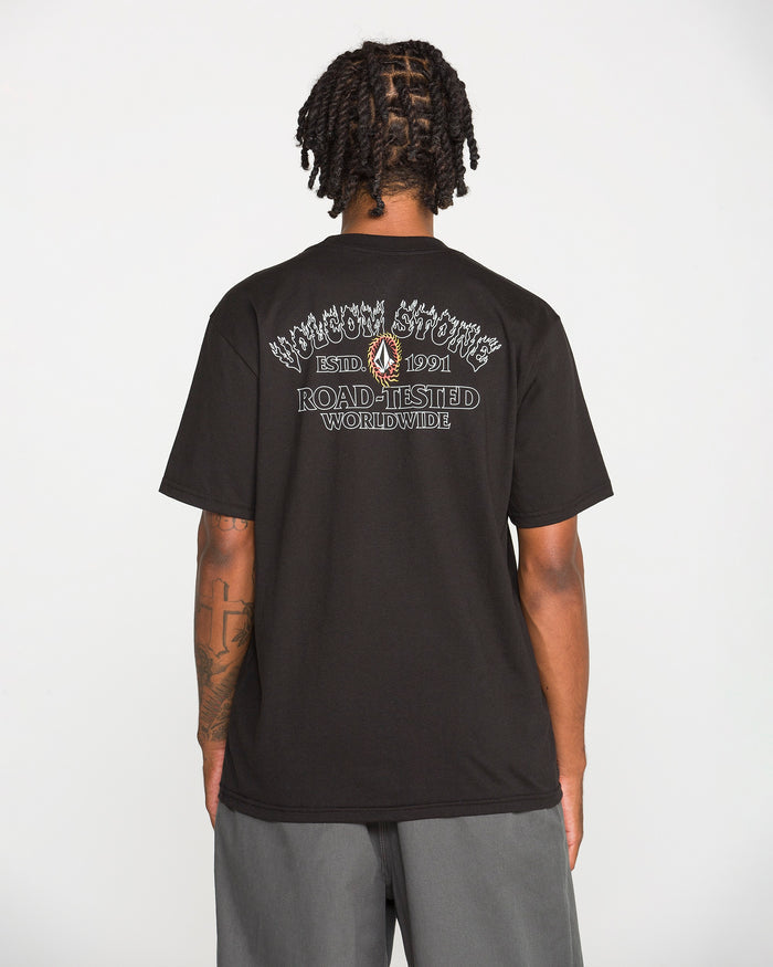 A person with short dreadlocks is seen from behind, wearing a black Volcom Men's Ringafire Short Sleeve Tee made of heavy-weight premium cotton, featuring white graphic text that reads “KINGDOM SPORTING ESTD 1991 ROAD-TESTED WORLDWIDE.” They are also wearing gray shorts.