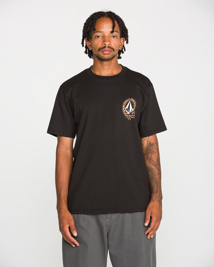 A person with shoulder-length dreadlocks wears the Volcom Men's Ringafire Short Sleeve Tee in classic black, crafted from heavy-weight premium cotton and featuring a small, colorful crest on the left chest. They pair it with gray pants and stand against a plain white background.