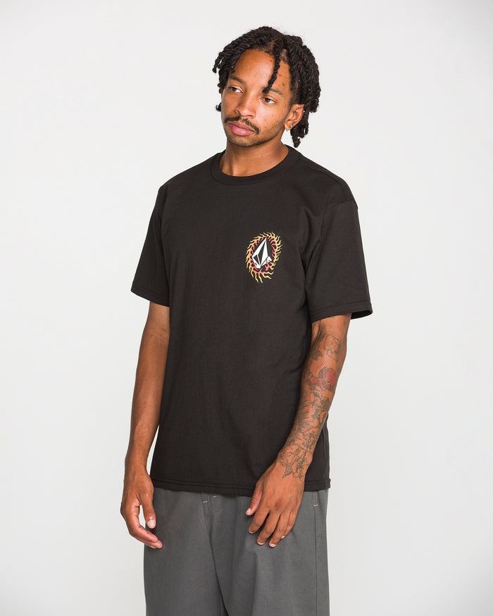 A man with tattoos and dreadlocks stands against a plain white background, wearing the Volcom Men's Ringafire Short Sleeve Tee—a classic fit black t-shirt made from heavy-weight premium cotton, featuring a small, colorful embroidered design on the left chest—paired with gray pants.
