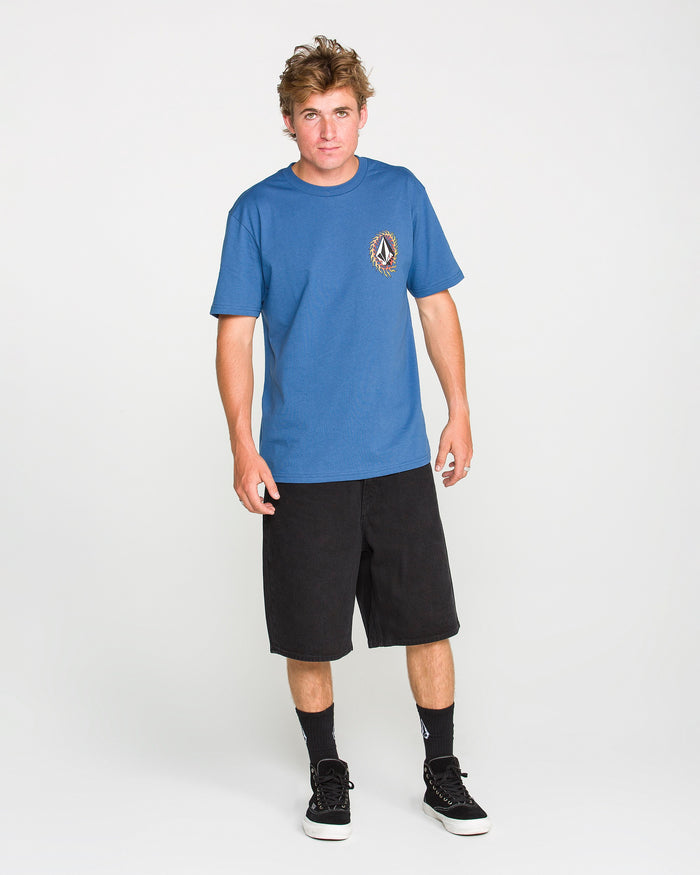 A person stands against a plain white background, wearing the Volcom Men's Ringafire Short Sleeve Tee—a blue, heavy-weight t-shirt crafted from premium ring-spun cotton and featuring a logo on the chest—paired with black shorts, black socks, and black sneakers.