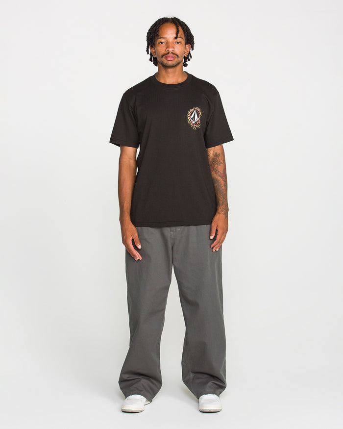 A person stands facing forward against a plain white background, wearing the Volcom Men's Ringafire Short Sleeve Tee in black—crafted from heavy-weight premium cotton with a small chest graphic—paired with gray wide-leg pants and white sneakers.