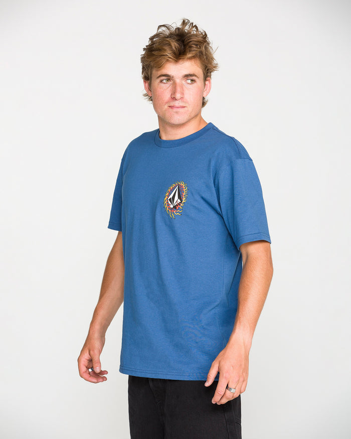 A person with tousled hair, wearing the Volcom Men's Ringafire Short Sleeve Tee in blue—crafted from premium ring spun cotton and featuring a colorful chest logo—stands turned slightly to the side against a plain white background.