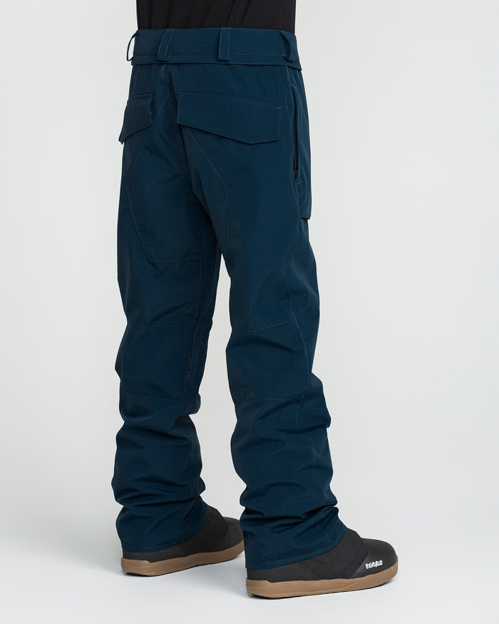 A man wearing Volcom Men’s Roan 20K dark blue snow pants and black snow boots is shown from behind against a plain white background.