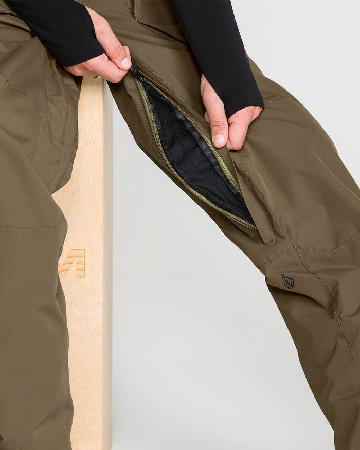 Wearing Volcom’s Men’s Roan 20K Pants in olive green—crafted from recycled fabric—a person unzips a thigh vent while seated on a light wooden bench.