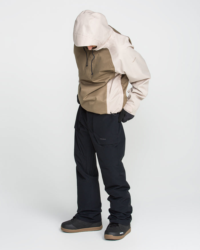 A person wearing a beige and brown hooded jacket, black pants, and dark shoes stands against a plain white background, looking down with hands in pockets. The outfit is completed with Volcom Men’s Roan 20K Pants—breathable ski pants crafted from recycled fabric snow pants—offering comfort and sustainability for outdoor adventures.