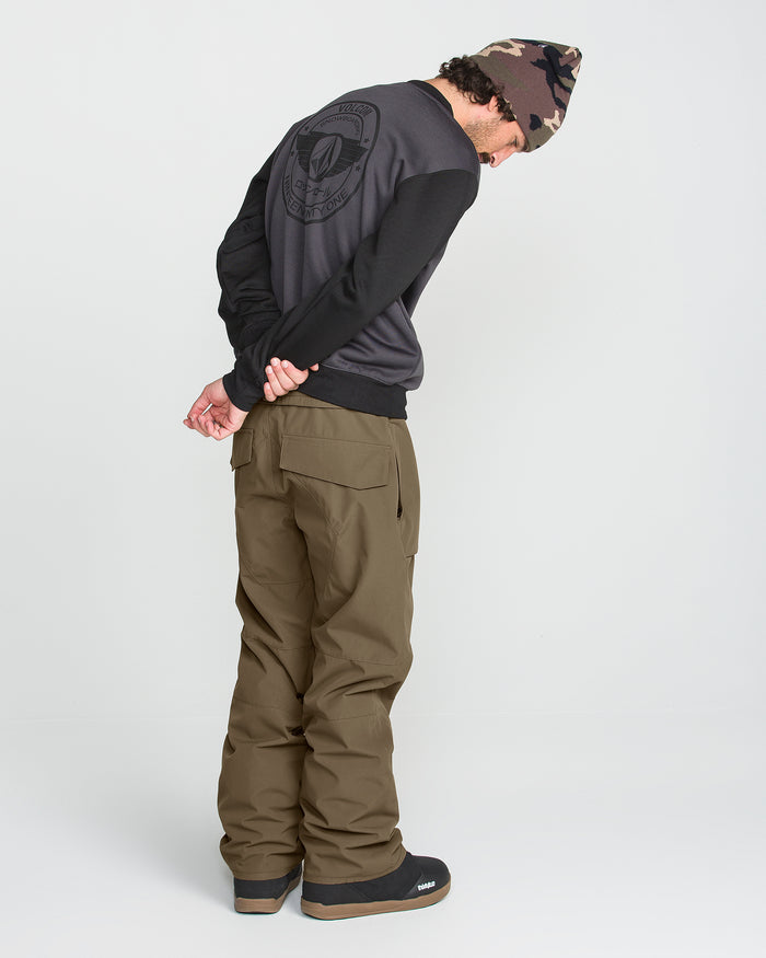 A person wearing a camo beanie, dark sweatshirt, and Volcom Men’s Roan 20K Pants—crafted from recycled fabric in olive—stands with hands behind their back, head bowed, facing away from the camera.