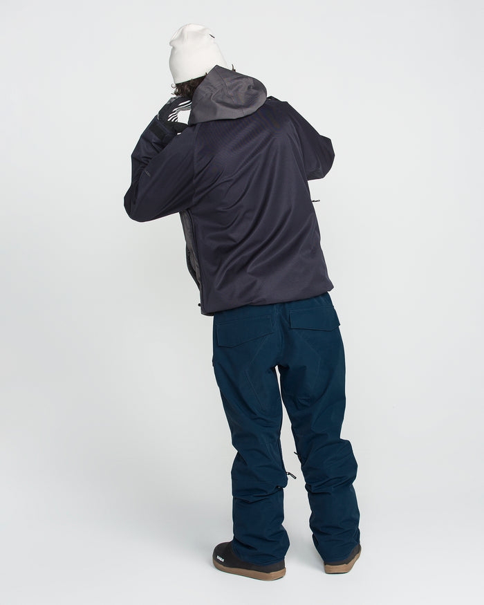 A person in a dark jacket, Volcom Men’s Roan 20K Pants in blue—crafted from recycled fabric—brown shoes, gloves, and a white beanie stands facing away, slightly turned, with one hand raised to their face against a plain white background.