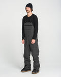 A man wearing a black beanie, a black long-sleeve shirt, and dark gray Volcom Men’s Roan Bib overalls stands facing slightly left against a plain white background. The stylish Volcom Men’s Roan Bib is reminiscent of recycled snow pants, seamlessly blending fashion with function.
